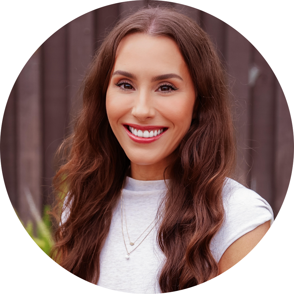 Portrait of a smiling woman with long wavy brown hair wearing a light gray t-shirt, outdoors against a wooden fence.