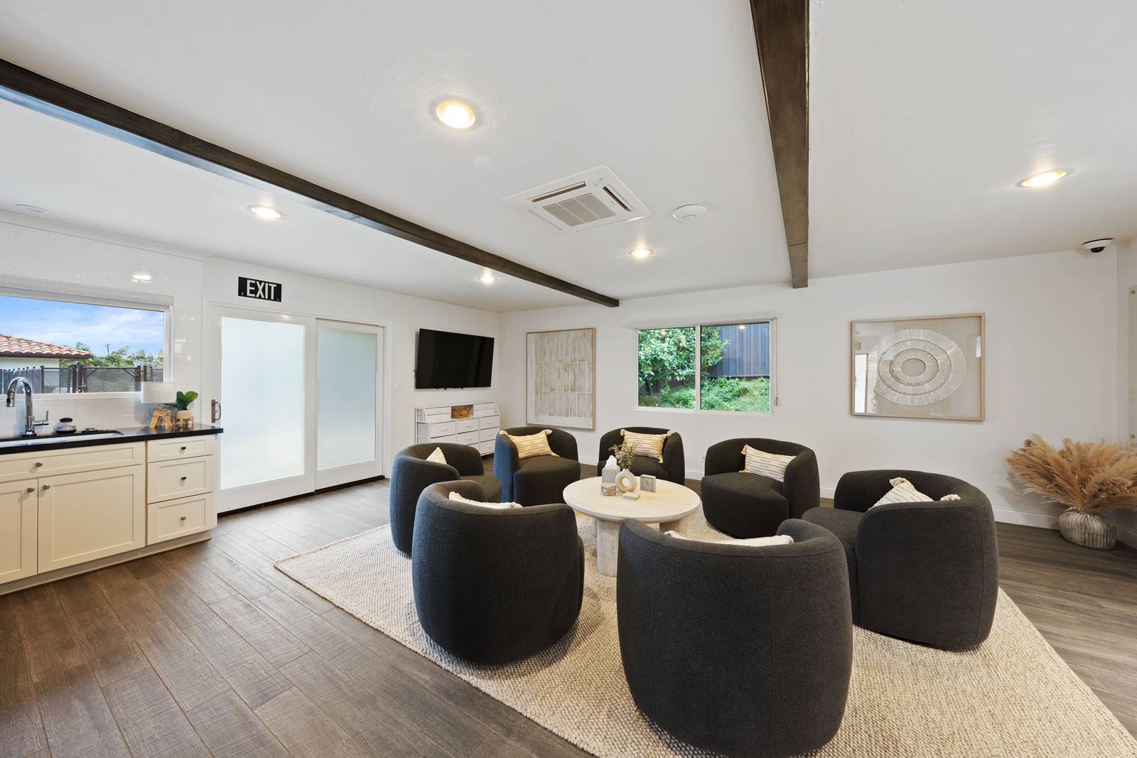 Open lounge area with eight dark gray barrel chairs around a round light-colored coffee table on a beige rug, bright and airy with white walls.