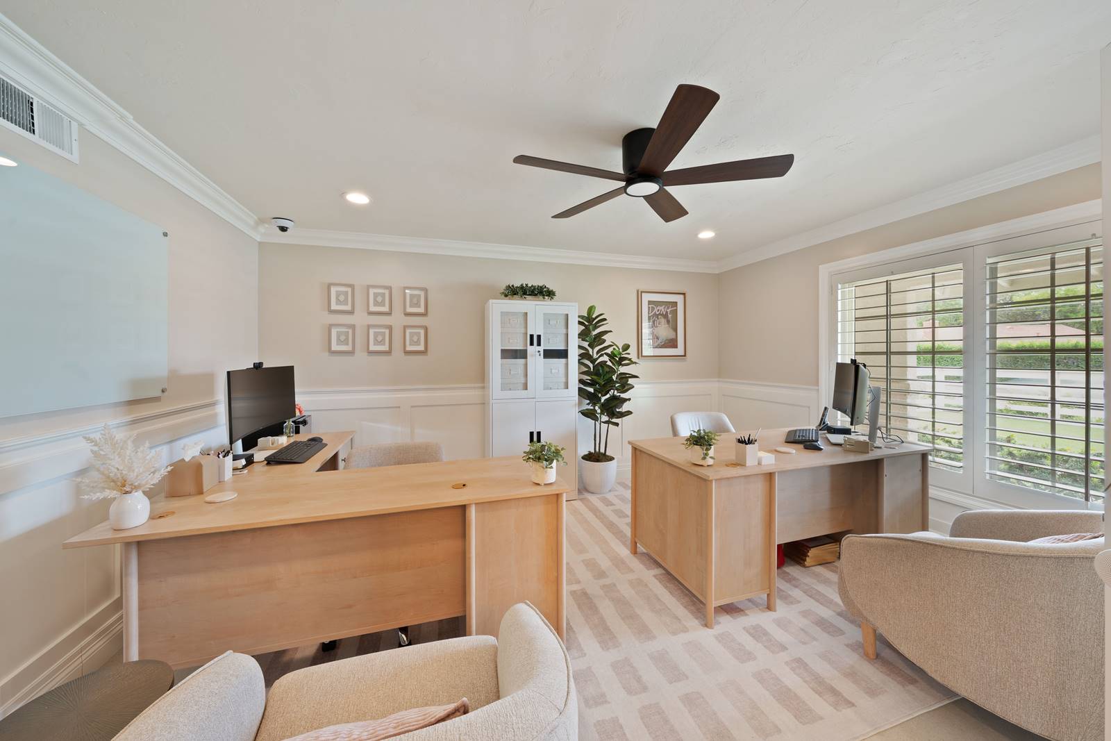 Bright, modern home office with two wooden desks, computers, and neutral seating under a ceiling fan. A large window with blinds lets in natural light.