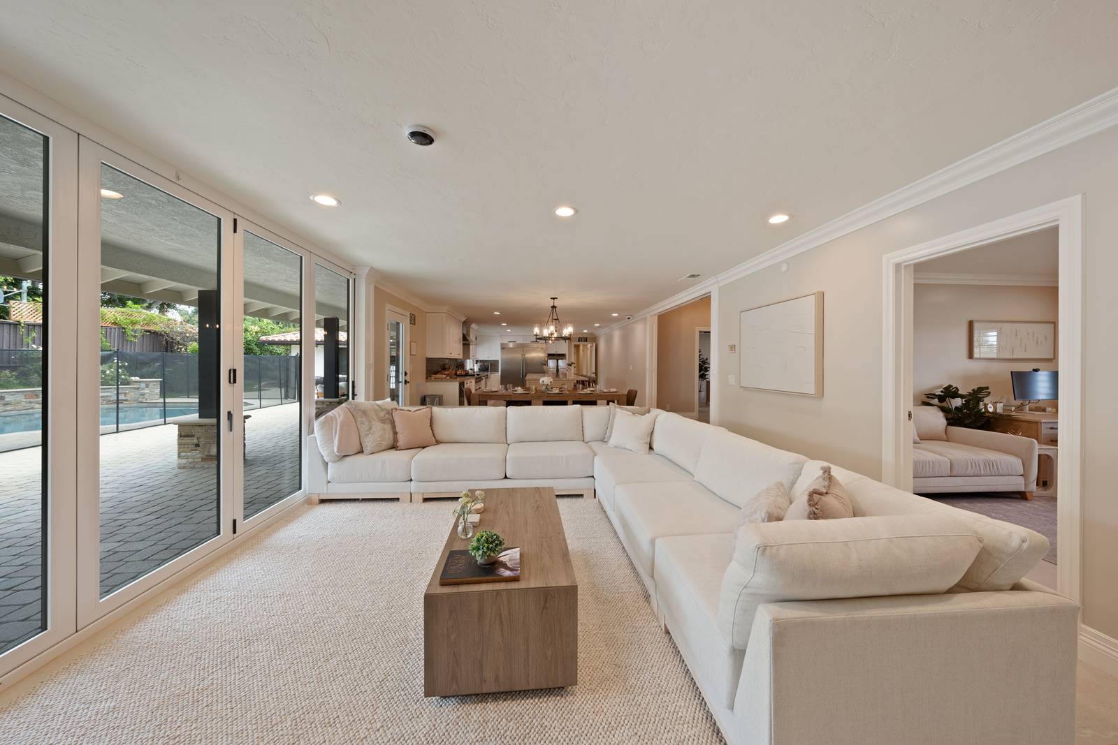 Open-concept living room with a large white sectional sofa, neutral decor, and glass sliding doors opening to a backyard pool area.