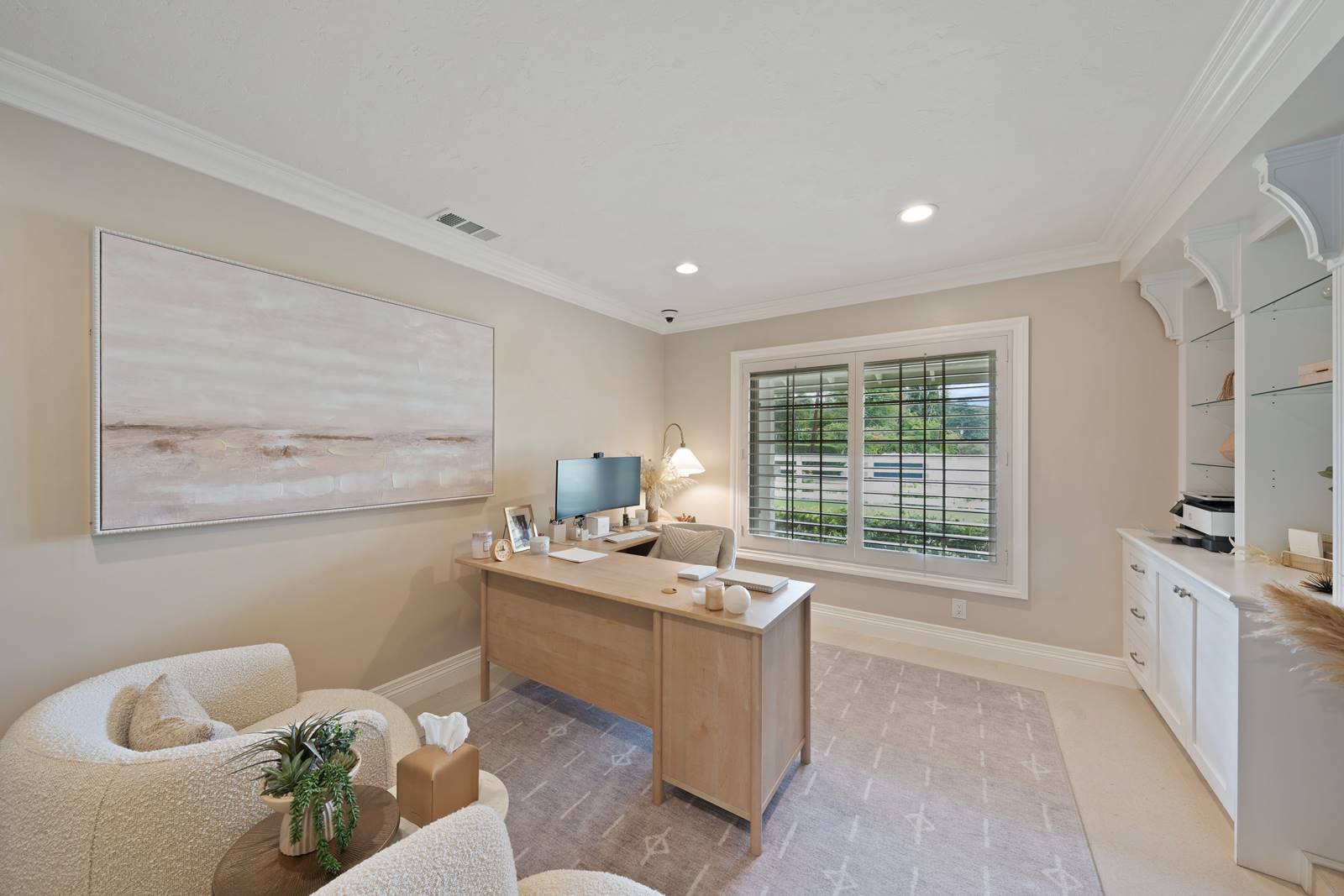 Cozy home office with a wooden desk facing a large window with white blinds and built-in shelves on the right.