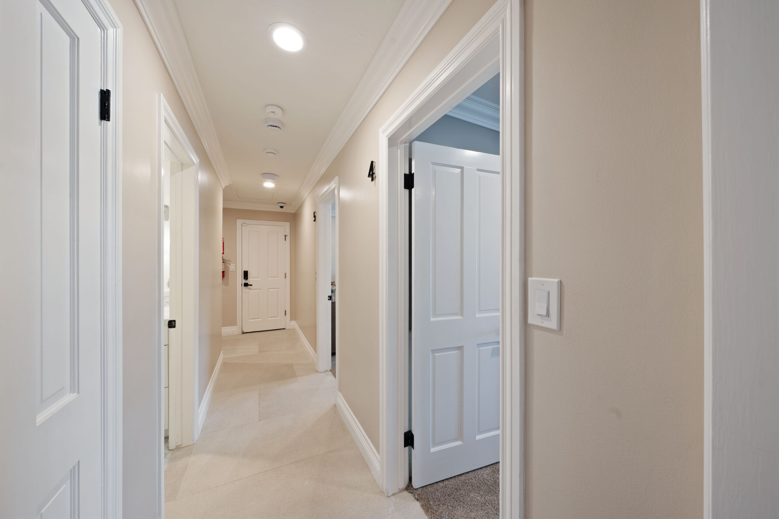 Interior hallway with white paneled doors on both sides and beige walls, lit by ceiling lights.