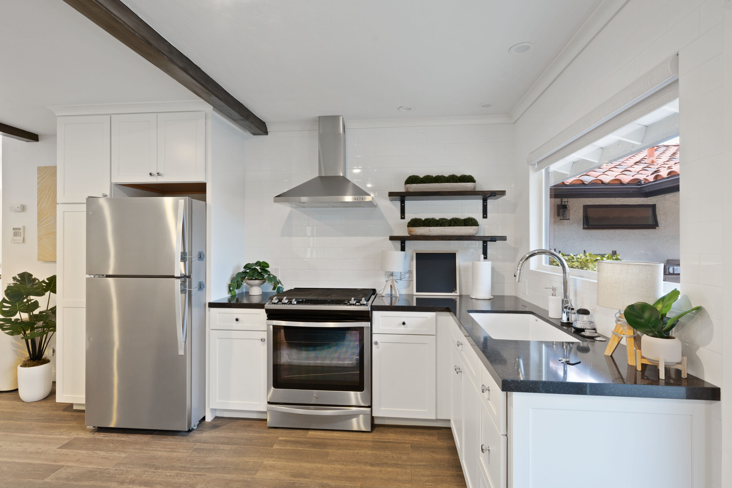 Bright modern kitchen with white cabinets, stainless steel fridge and stove, and floating dark-wood shelves with greenery near a large window.