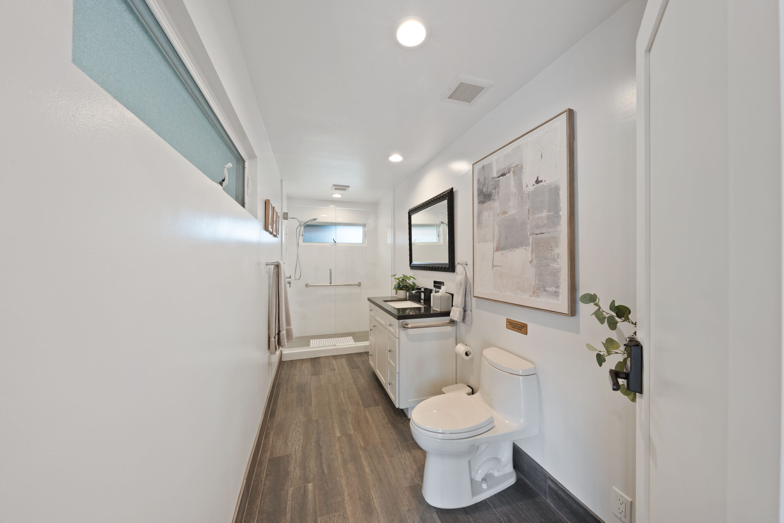 Narrow, modern bathroom with white walls, wood-look flooring, a white vanity and sink, and a large abstract painting above the counter.
