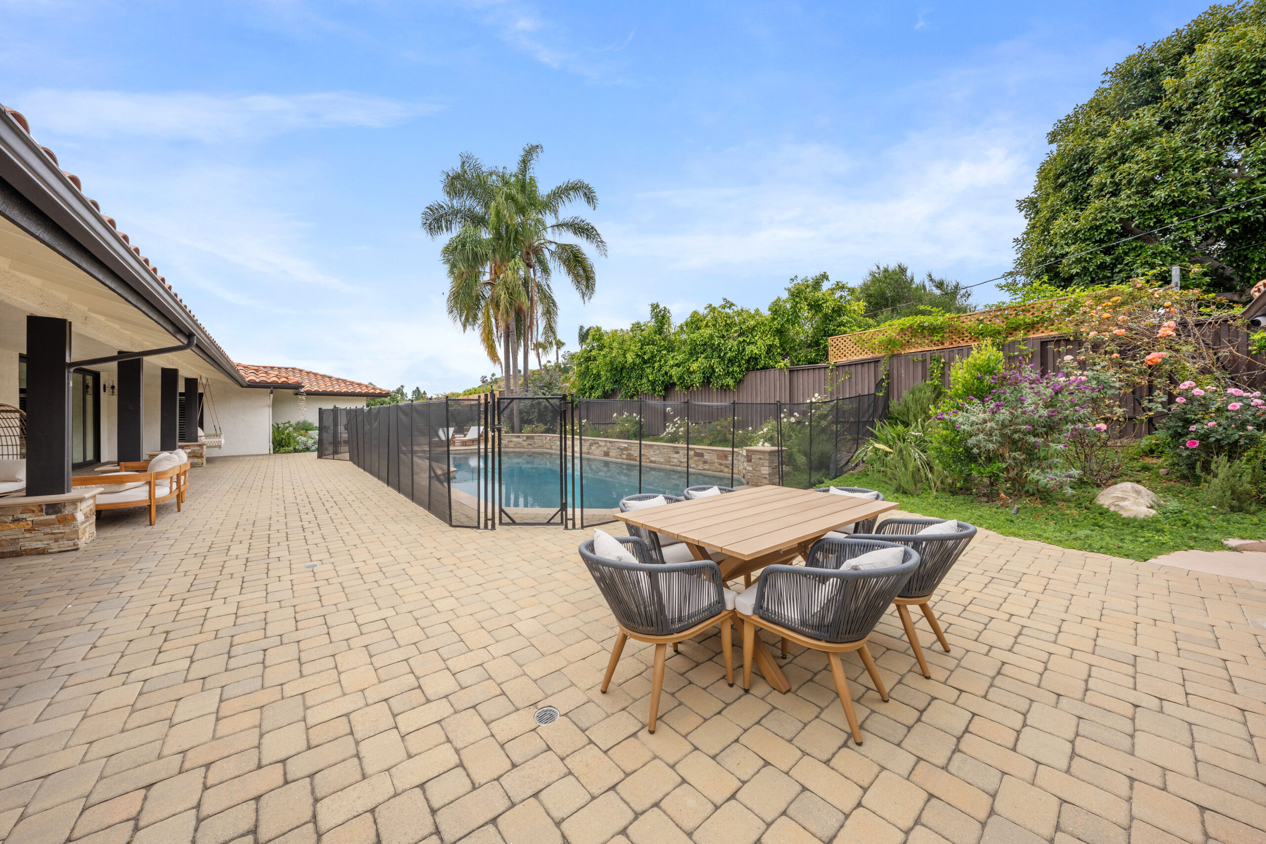 Backyard patio with a stone-tiled deck, round wooden table and black wicker chairs near a fenced rectangular pool and palm trees in the background.