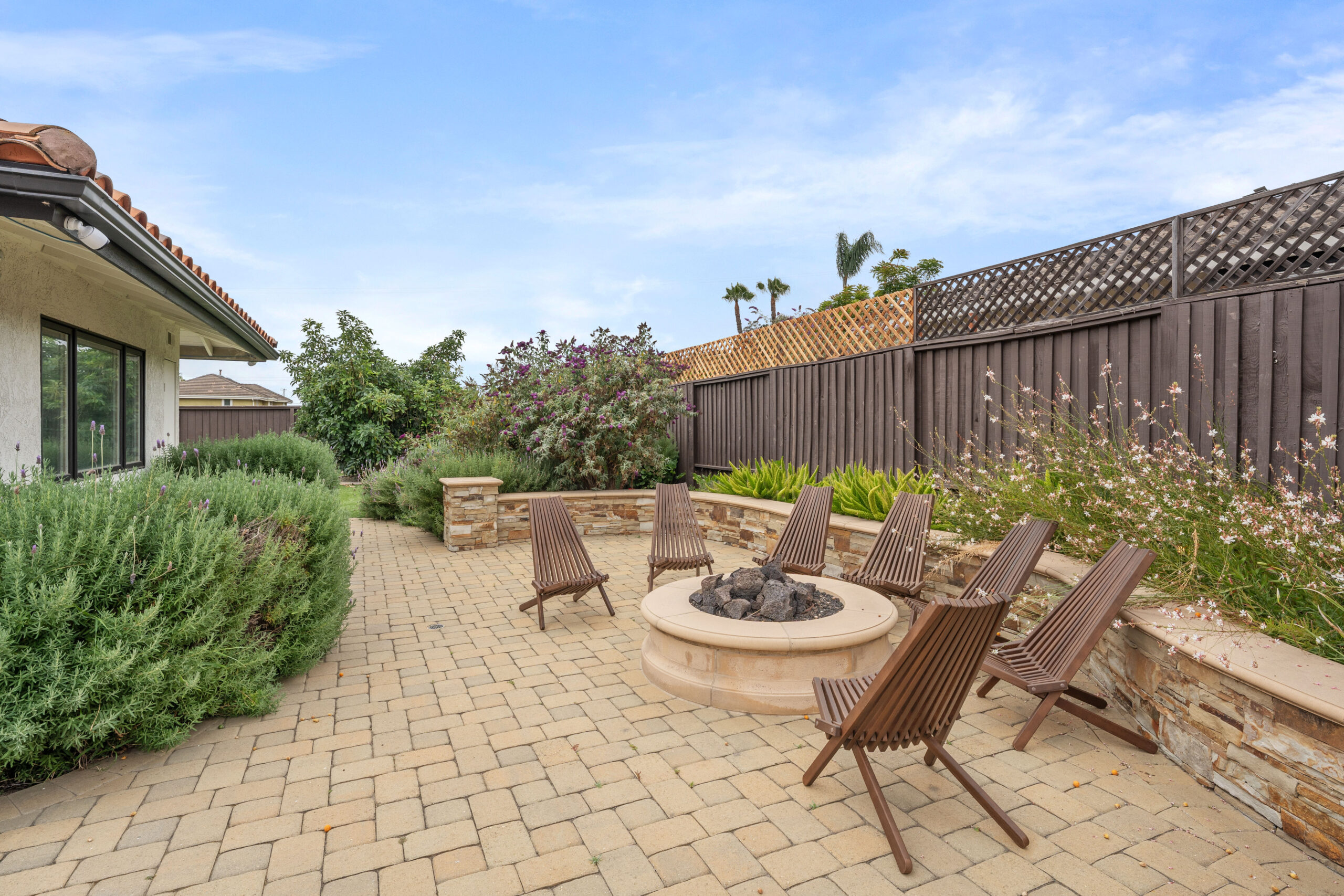 Backyard patio with a circular stone fire pit and brown wooden chairs on a brick-paved surface, surrounded by shrubs and a tall wooden fence.
