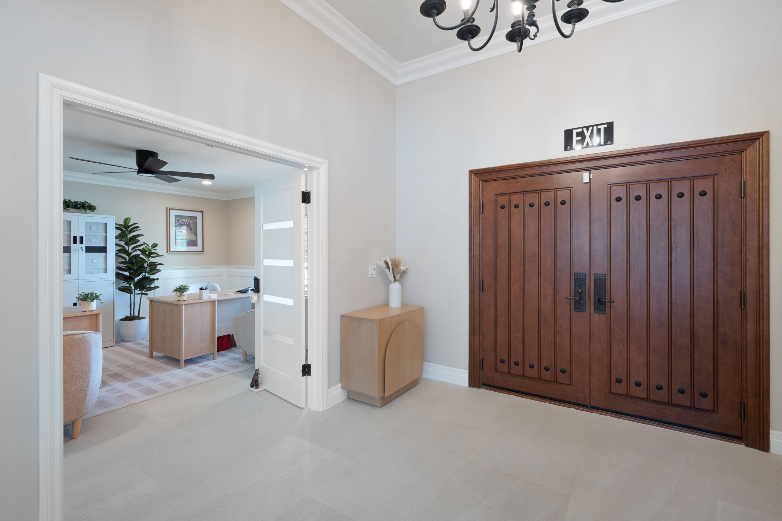 Foyer with large dark wood double doors, exit sign above, and a black chandelier in the ceiling; open doorway leads to an office with plants.