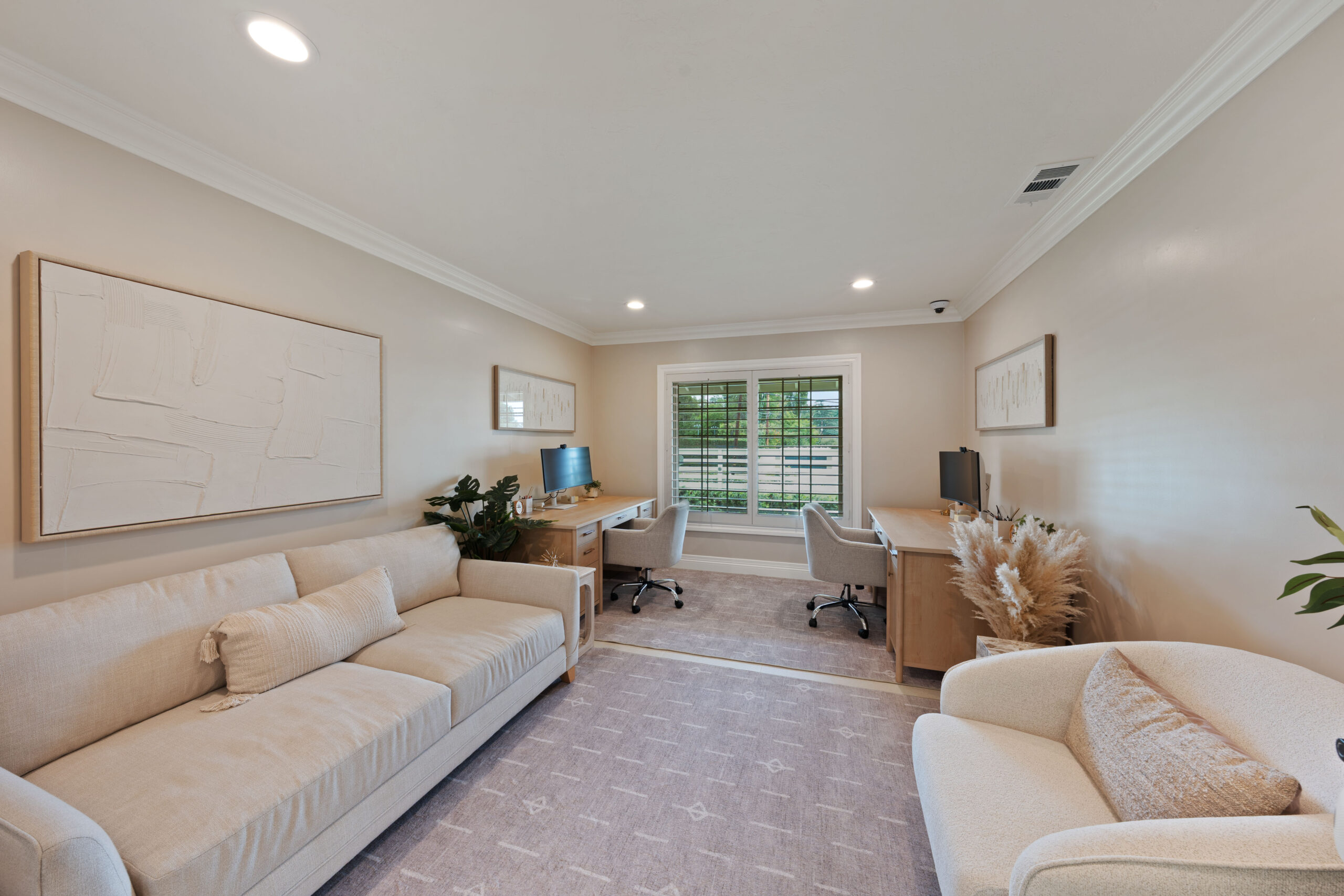 Bright home office living area with two desks, beige seating, and a large window with blinds; neutral decor and plants nearby.