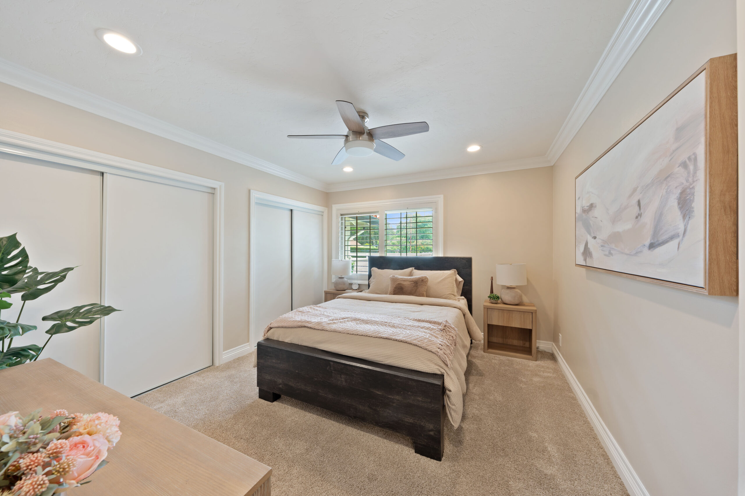 Bedroom with a dark wood platform bed, beige bedding, matching nightstands, and a ceiling fan in a bright, neutral-toned room with sliding closet doors and a large framed mural on the wall to the right.