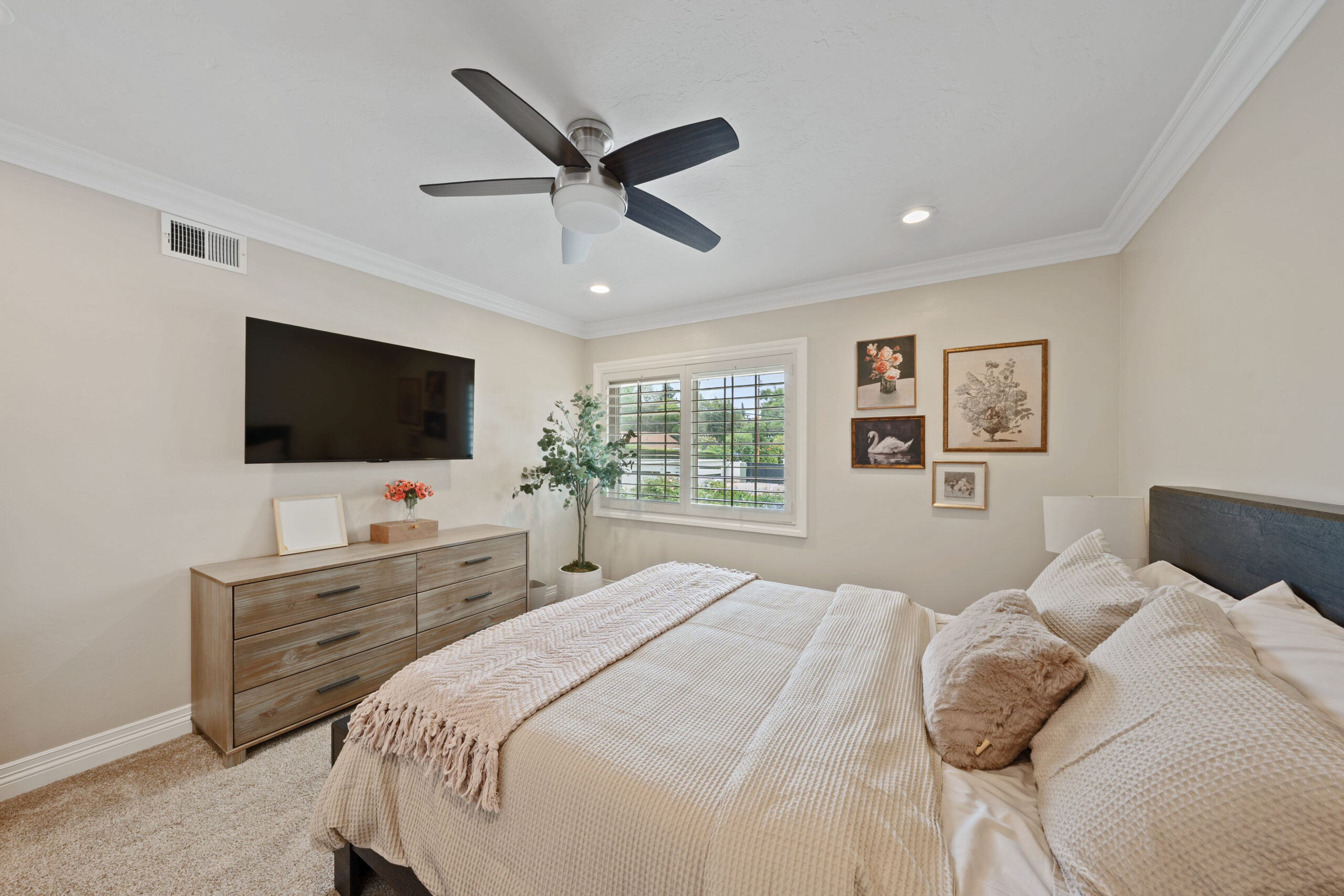 Bedroom with beige walls, ceiling fan, dresser, wall-mounted TV, window with blinds, potted plant, and framed artwork. Notable: cozy, modern decor with soft textiles on bed.