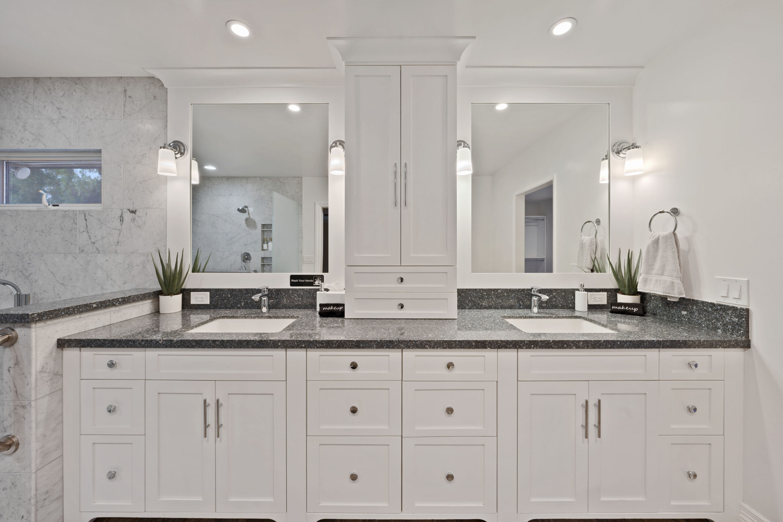Bright white double vanity with a granite countertop, two sinks, and a tall central storage cabinet flanked by mirrors in a modern bathroom.