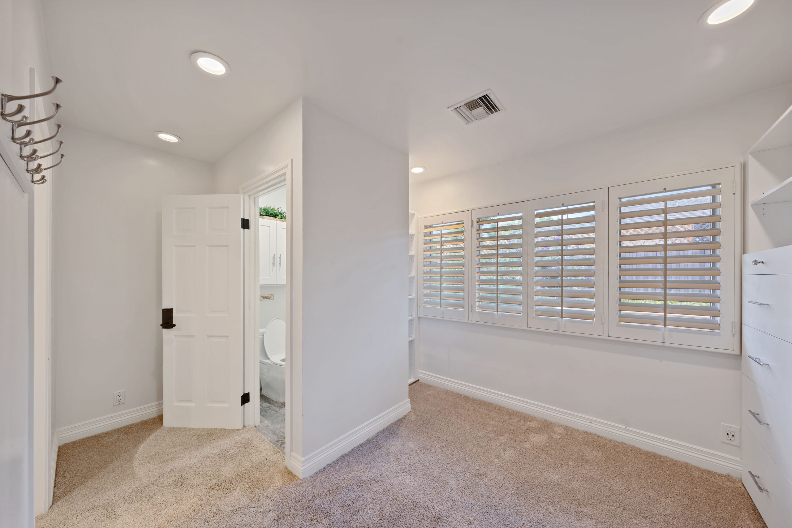 Bright laundry/mudroom with white cabinets on the right, light carpet, and row of shuttered windows along the far wall; doorway to an adjacent bathroom visible on the left.
