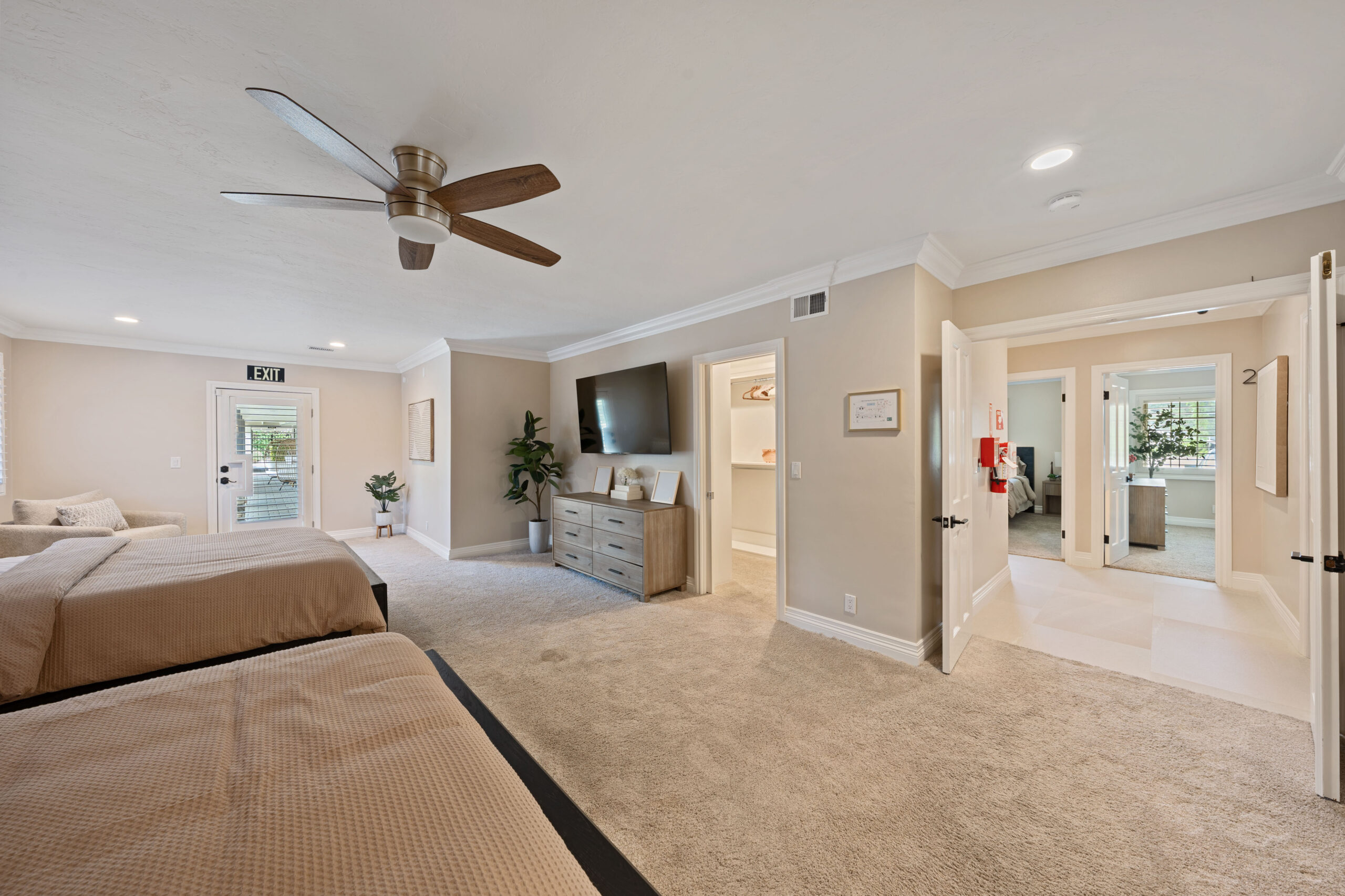 Bedroom with two beige-covered beds, a central ceiling fan, and a dresser with a wall-mounted TV across from the beds.