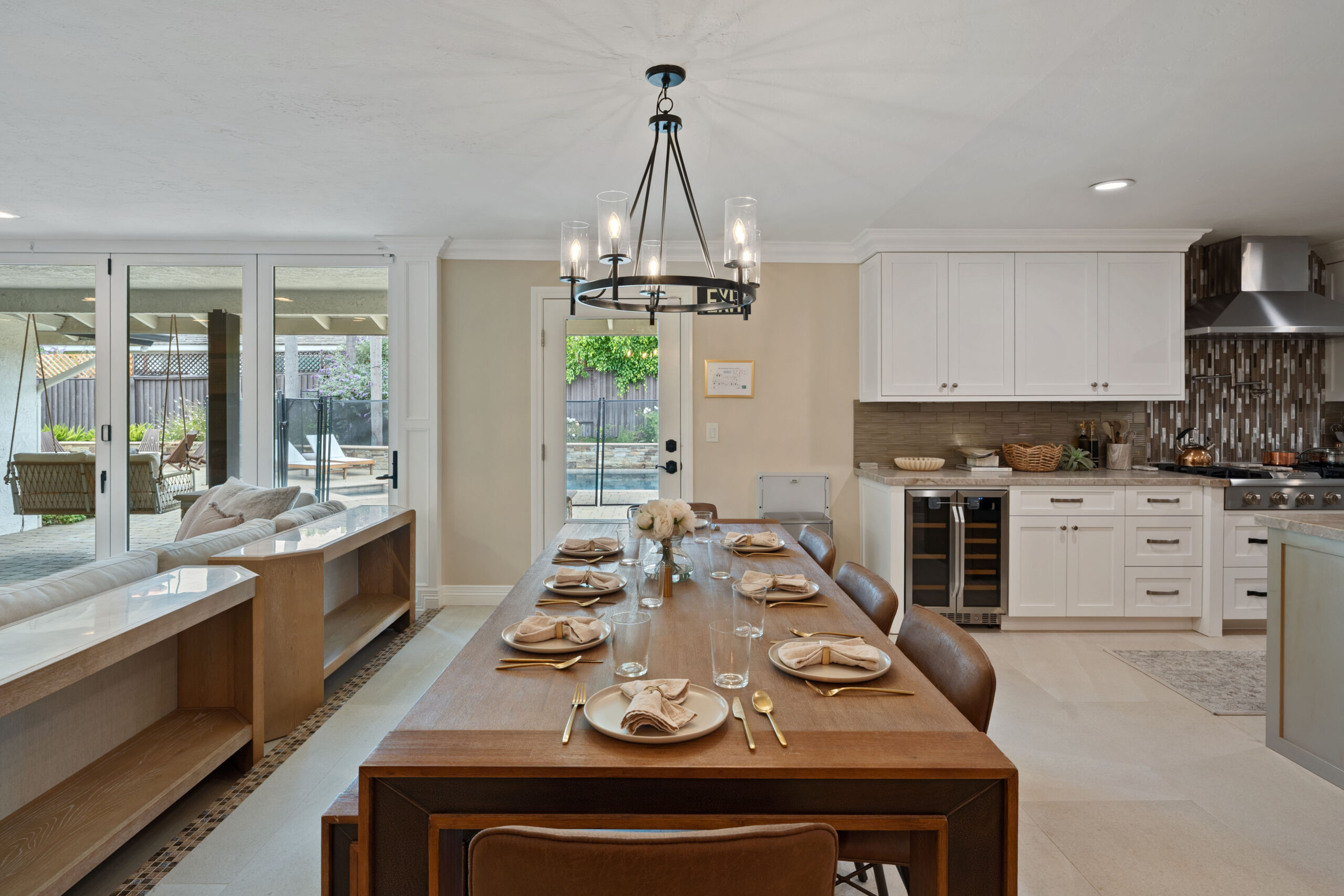 Long wooden dining table set for a meal in a bright open-concept kitchen and living area, with a wrought-iron chandelier overhead.