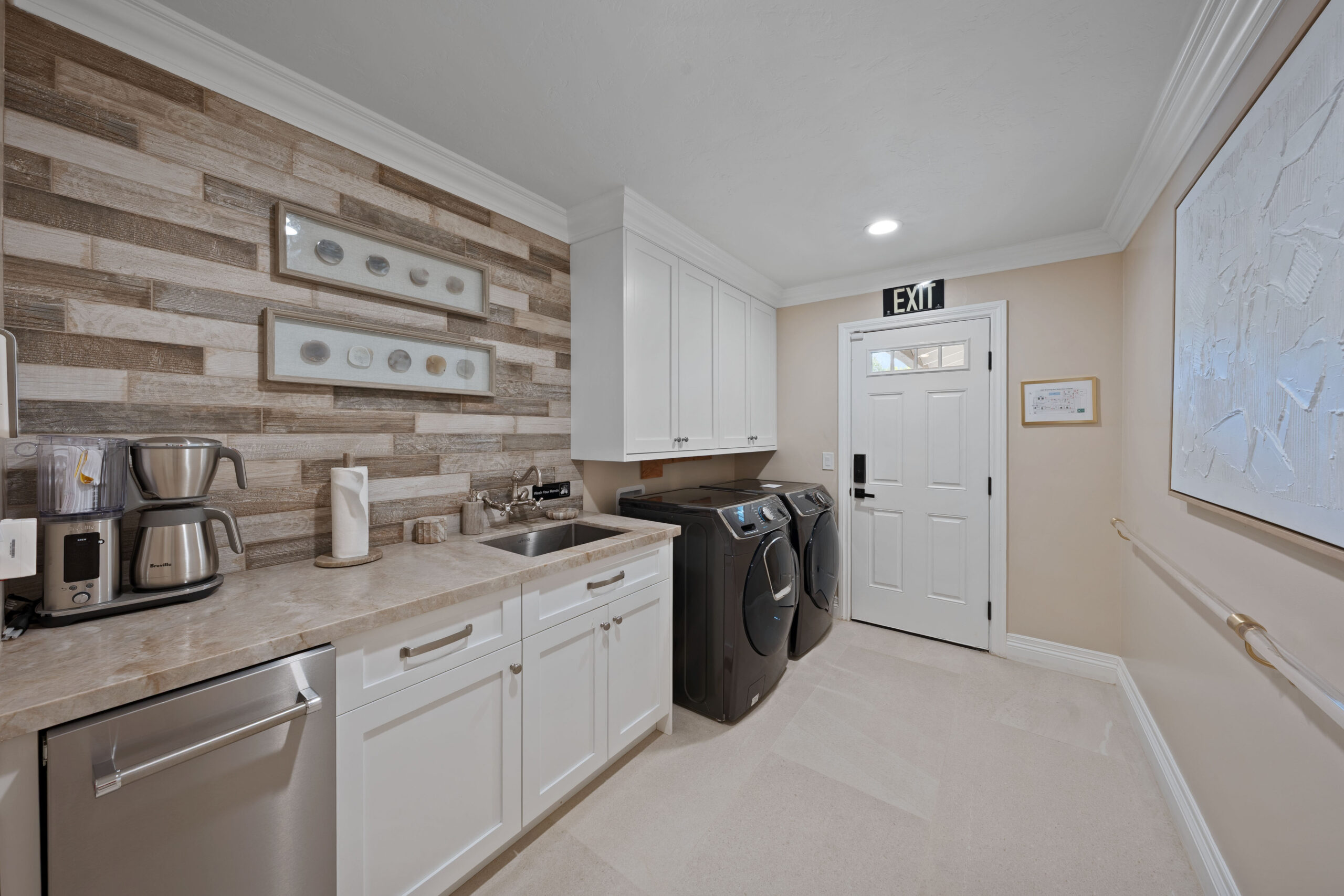 Laundry room with white cabinets, washer and dryer, stainless sink, and wood-plank accent wall plus framed shell displays on left wall.