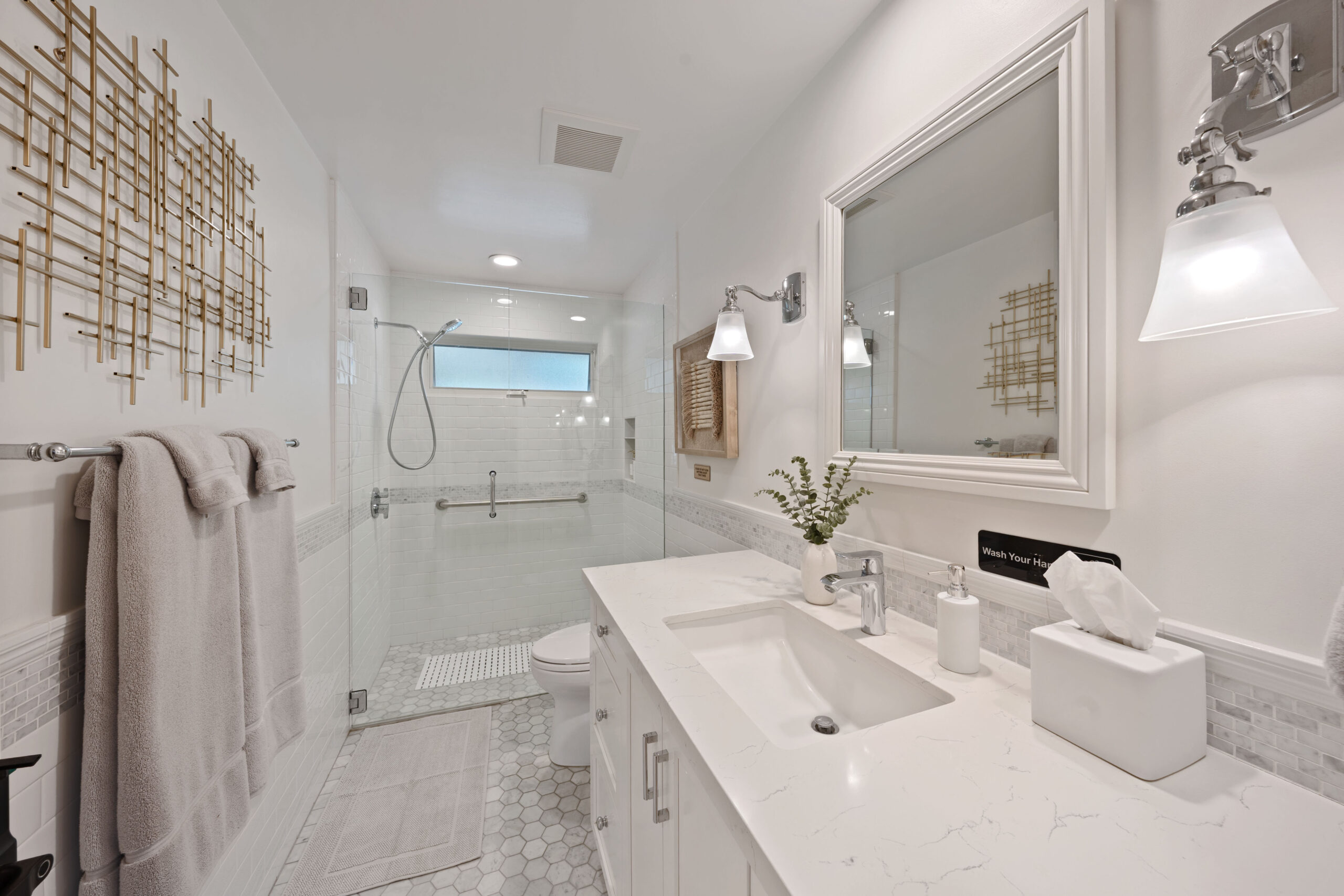 Modern white bathroom with a glass shower, marble vanity, and gold geometric wall art.